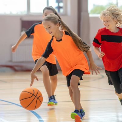 Children playing basketball indoors, wearing orange and red jerseys.