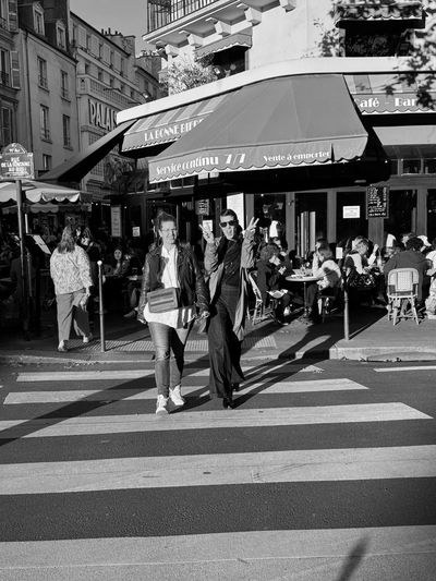 Two people crossing a street in front of a café with outdoor seating.