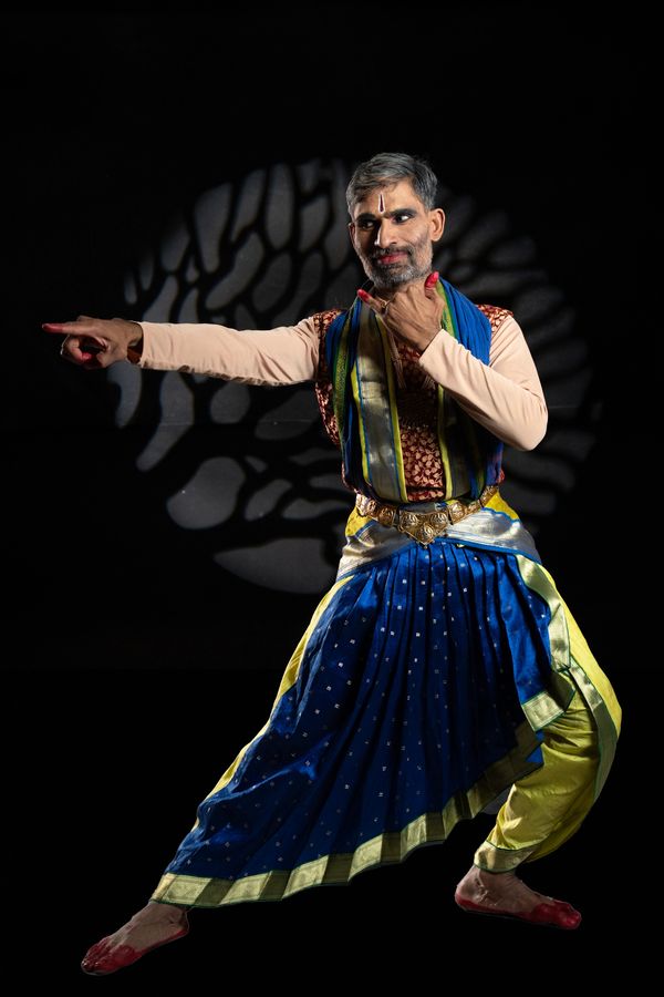 Male classical Indian dancer in vibrant attire poses against artistic backdrop.