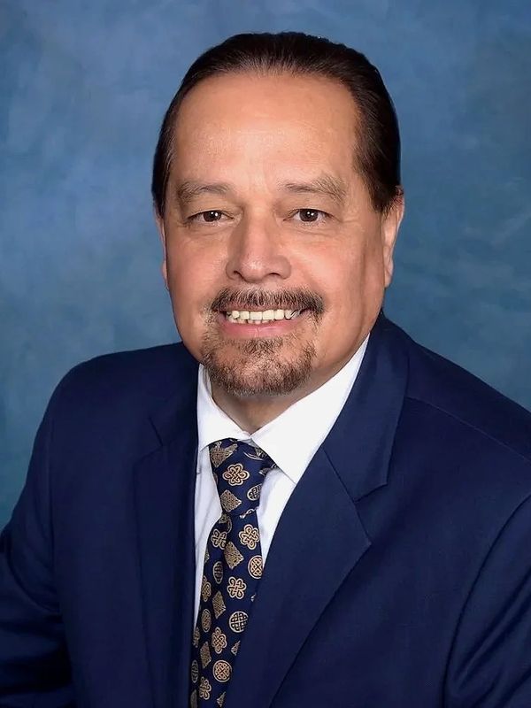 A man in a blue suit and patterned tie smiling against a blue background.