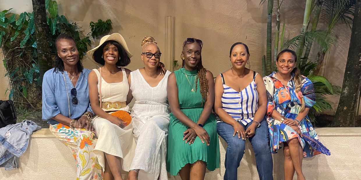 Six women sitting together outdoors, smiling and dressed in colorful summer outfits.