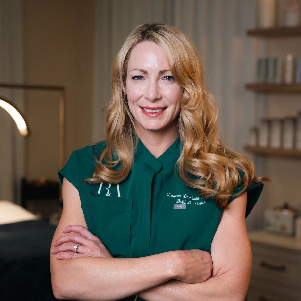Smiling blonde woman in green uniform stands with arms crossed in a professional setting.