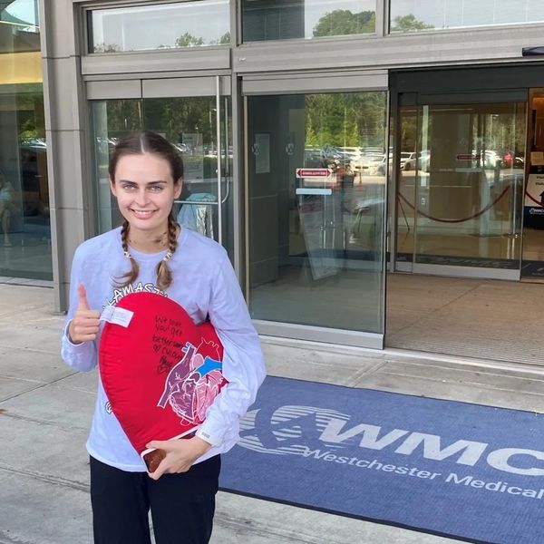 Young woman holding a heart-shaped pillow, standing outside a medical center.