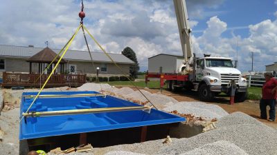 A crane is lowering a blue pool shell into a prepared ground site.