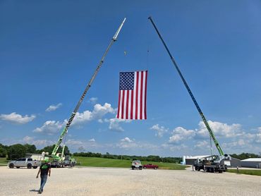 Two cranes hoisting a large American flag outdoors on a sunny day.