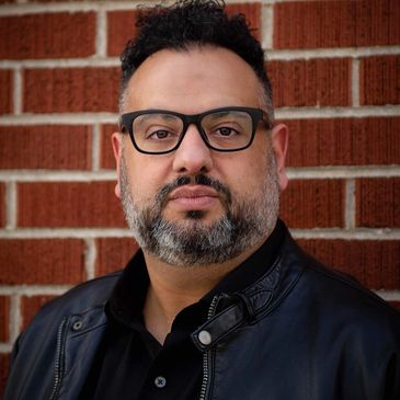 Man with glasses and beard against a brick wall background.