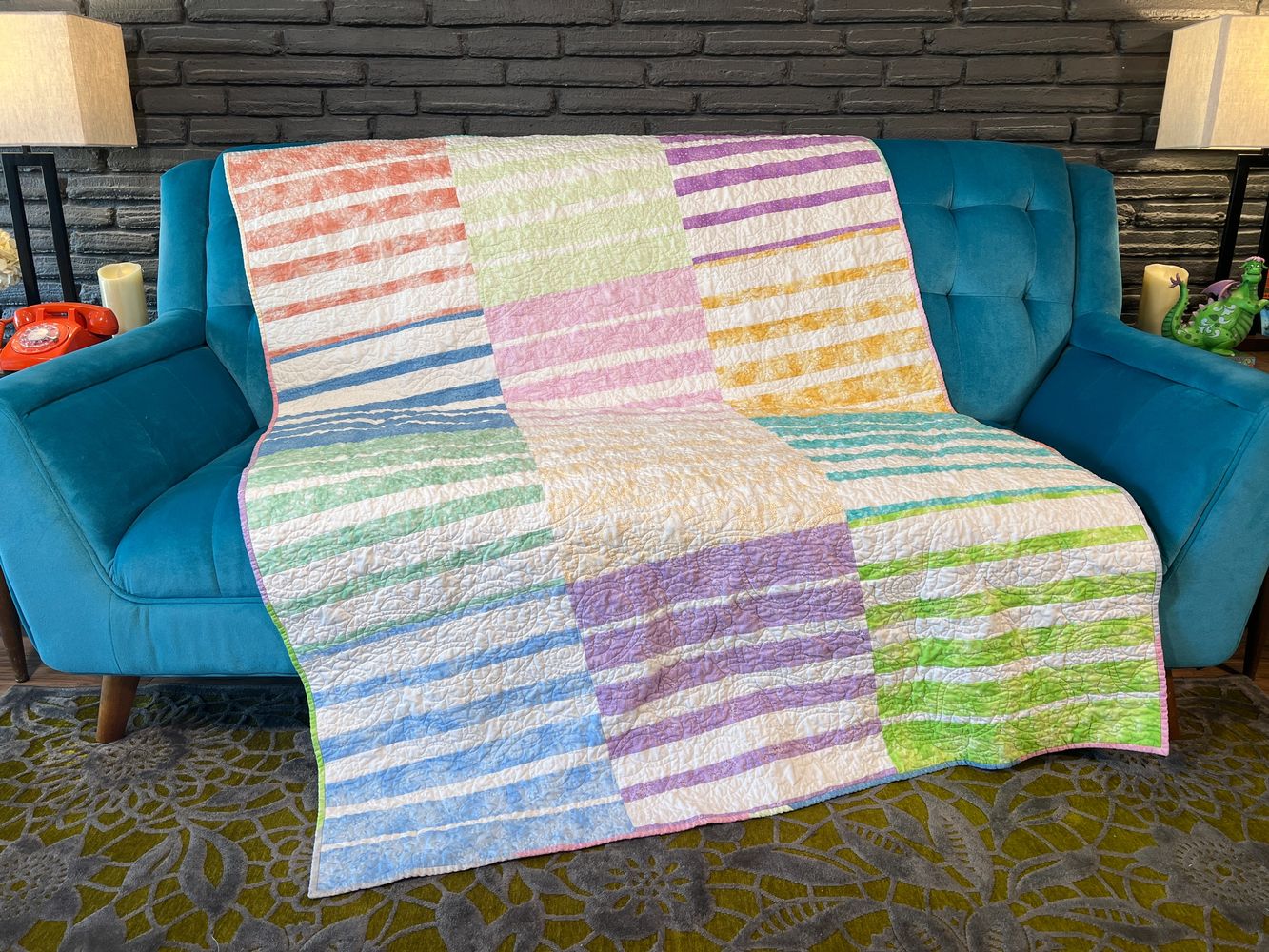 A  patchwork quilt with pastel stripes arranged in large blocks is draped over a mid-century sofa.