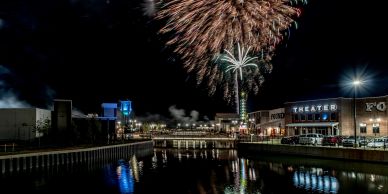 Fireworks burst over a river at night near a theater.