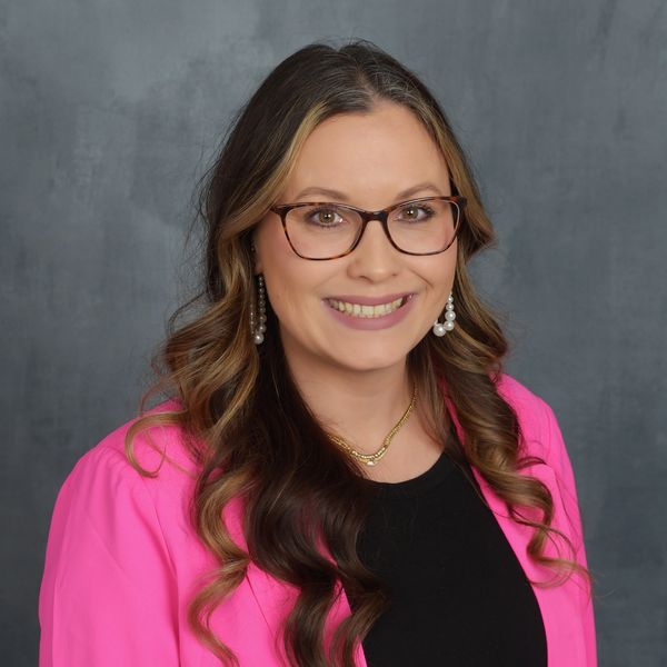 Woman with glasses and curled hair wearing a pink blazer and pearl earrings.