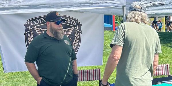 Two men talking at an outdoor event with a Coin Vets banner.