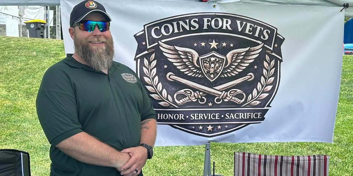 A man stands smiling in front of a 'Coins for Vets' banner at an outdoor event.