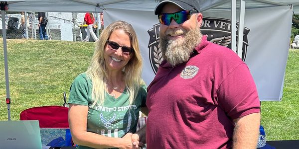 Two people shaking hands and smiling outdoors at a booth.