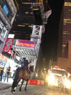 Mounted police officer patrols a busy urban street at night with illuminated signs and tall buildings.