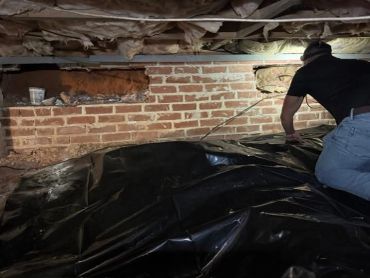 Man working in a dimly lit crawl space with black plastic covering.