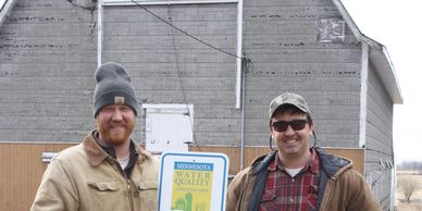 Two men stand outside holding a Minnesota water quality sign, dressed warmly in jackets and hats.