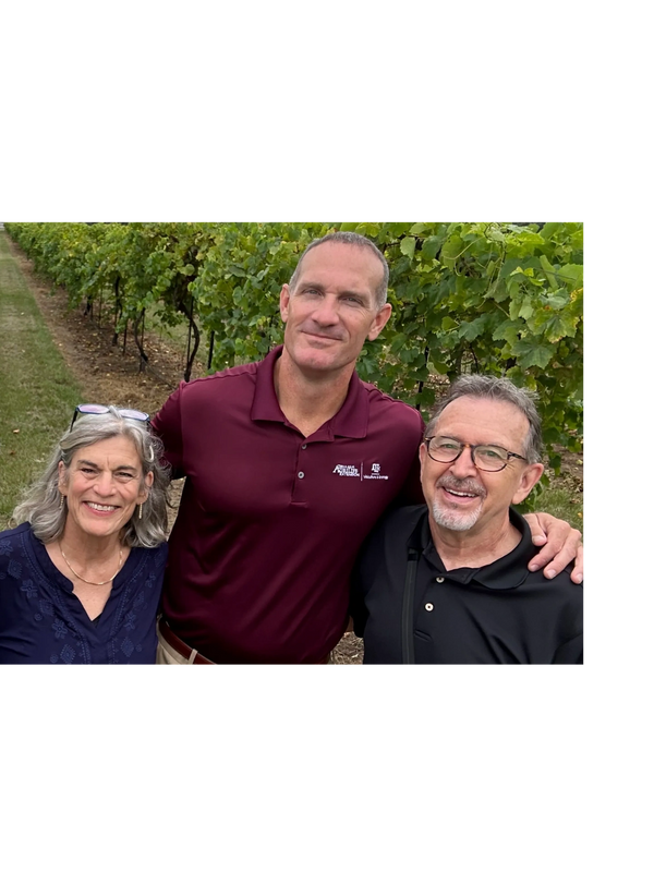 Three people smiling together in a vineyard with lush green vines behind them.