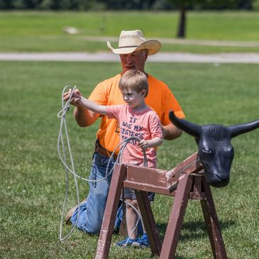 A child learning to lasso a practice bull with an instructor in a field.