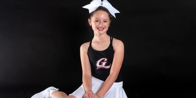Young cheerleader in black top and white skirt with bow smiling.