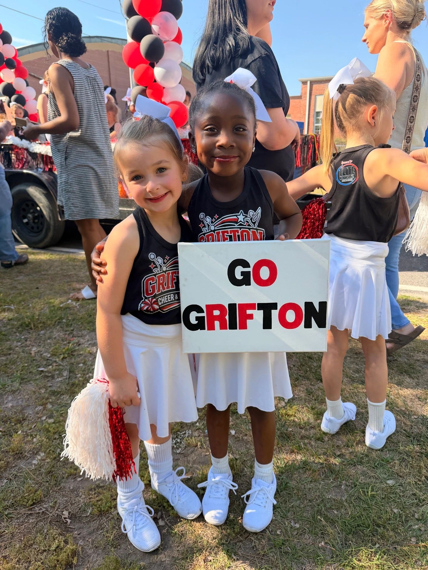 Two young cheerleaders smile holding a 'Go Grifton' sign at an outdoor event.