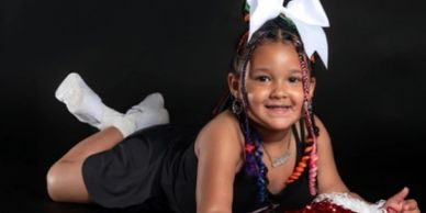 Smiling girl lying on her stomach with colorful braids and a big white bow.