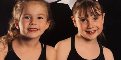 Two young girls smiling and posing in black tank tops against a dark background.