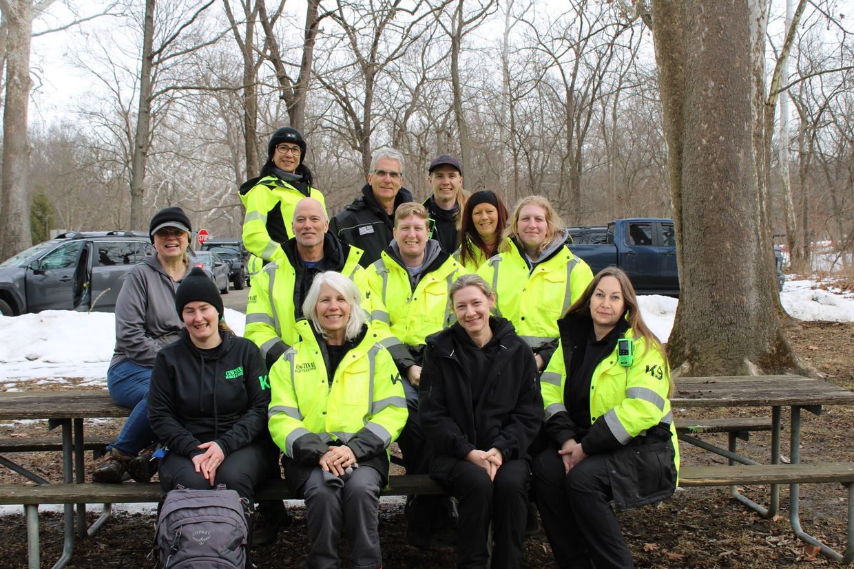 Group of people in high-visibility jackets sitting on picnic tables outdoors in winter.
