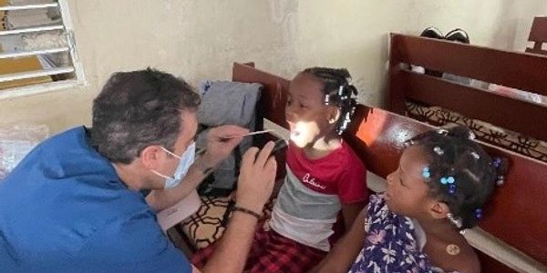 Doctor examining a child's throat while another child watches.