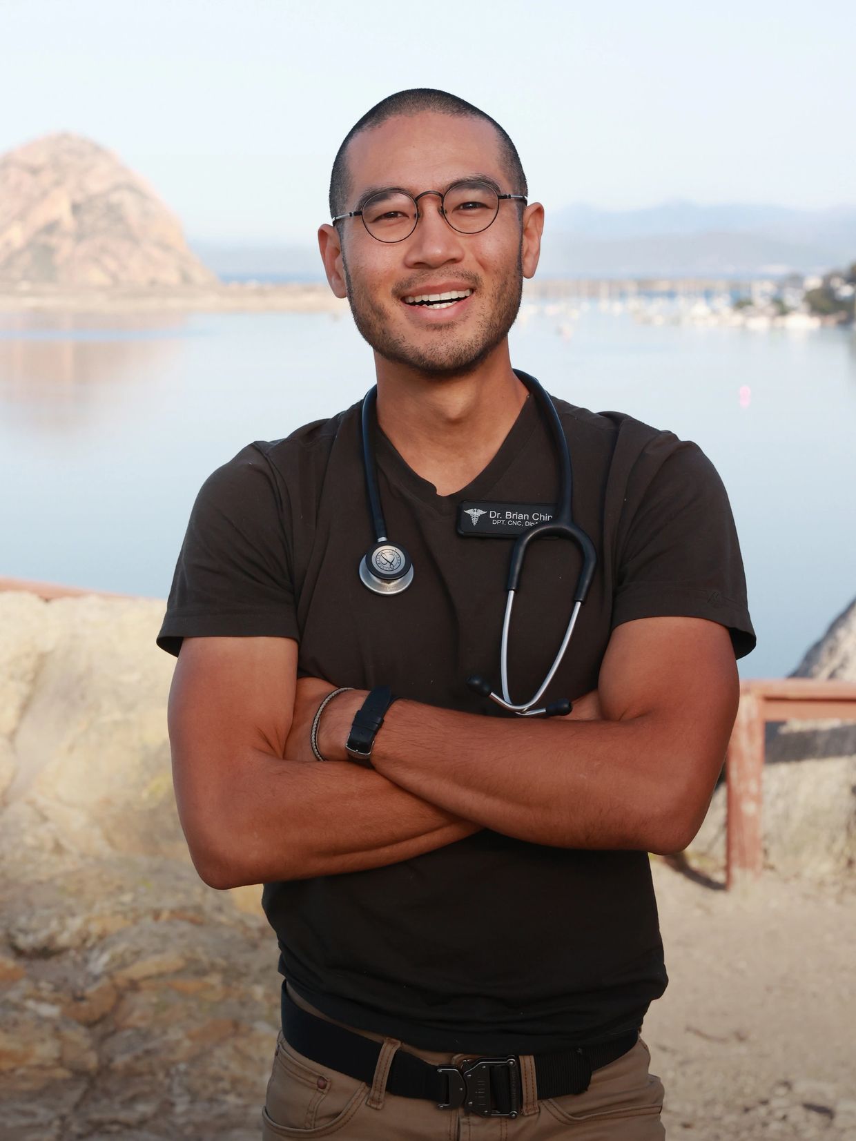 Smiling doctor with glasses and stethoscope outdoors by water and mountains.