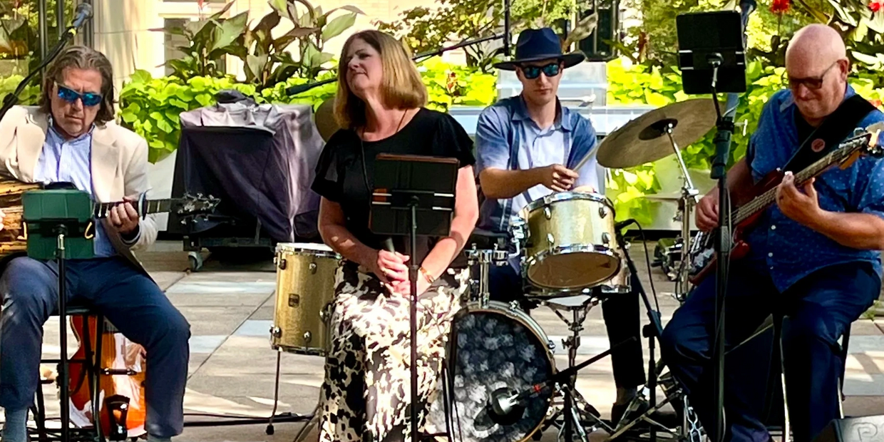 A band of four musicians performing outdoors with guitars, drums, and a vocalist.