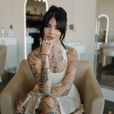 Stylist with dark hair and full tattoos posing in salon chair, wearing white dress