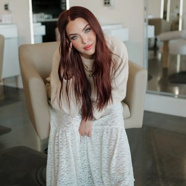 Stylist with long auburn hair in cream sweater and lace skirt sitting in salon chair