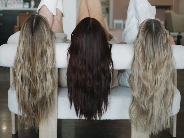 Three women with long blonde and brunette hair extensions draped over a white chair in a salon.
