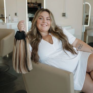 Stylist with long wavy brunette hair in white dress holding hair extension swatches in salon chair