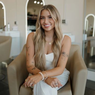 Stylist with long blonde hair sitting in salon chair, smiling in white top and striped pants.