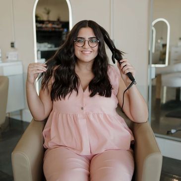 Stylist with dark wavy hair and glasses curling hair with iron, wearing pink outfit.