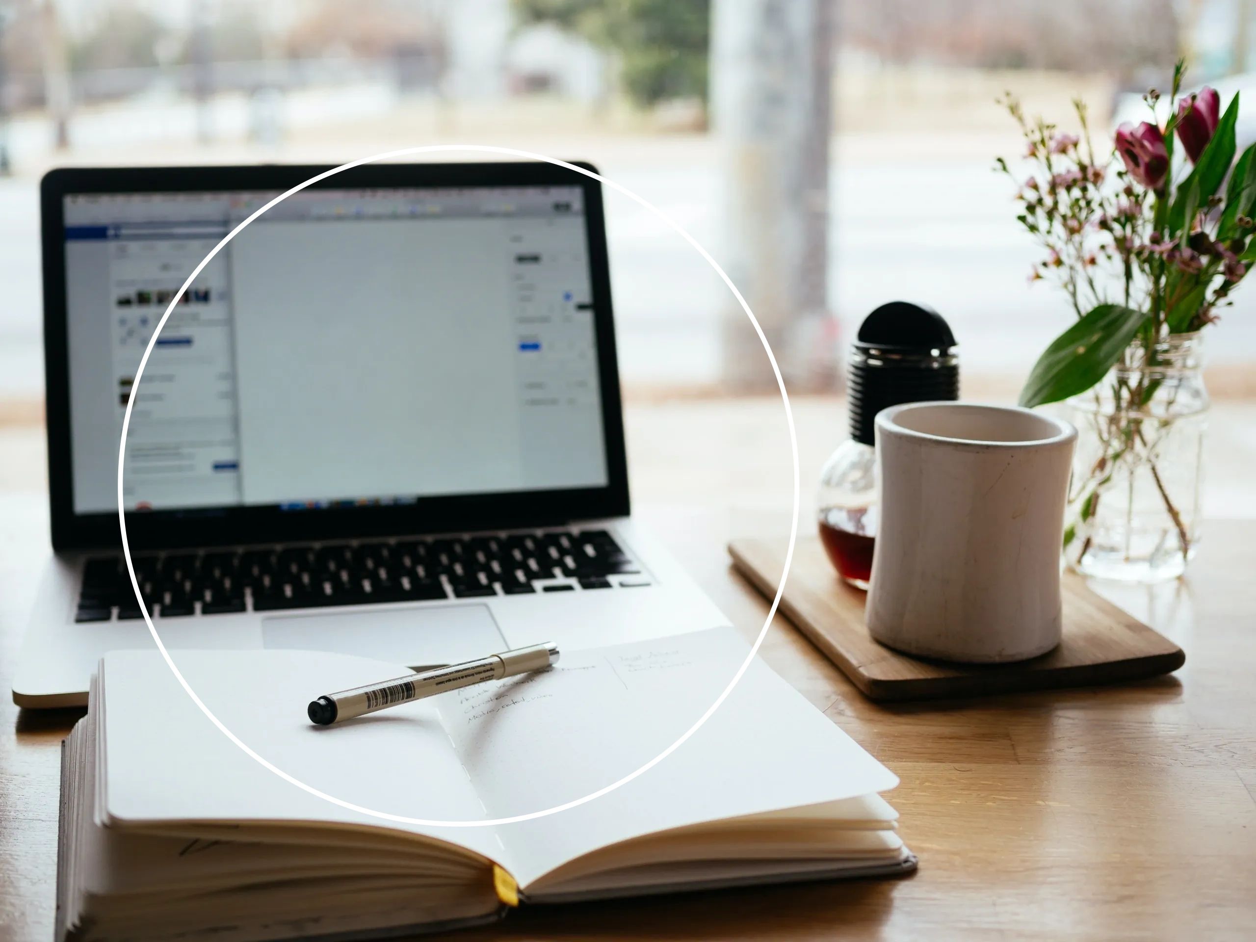 Open notebook and pen on desk with laptop, coffee, and flowers in cozy workspace.