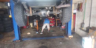 Mechanic underneath a car on the lift doing suspension work.  The mechanic is wearing tan shorts and a blue shirt.  There are tools on the ground and tires piled up on the left