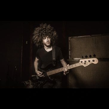 A man with a cool hairstyle plays a bass guitar in a dark room.
