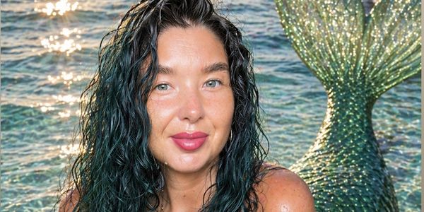 Woman with wet hair posing in front of a sparkling mermaid tail by the water.
