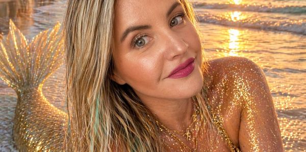 Woman with glittery gold mermaid tail poses on the beach at sunset.