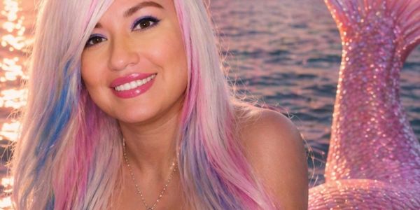 Smiling woman with pastel pink and blue hair near a sparkling mermaid tail by the water.