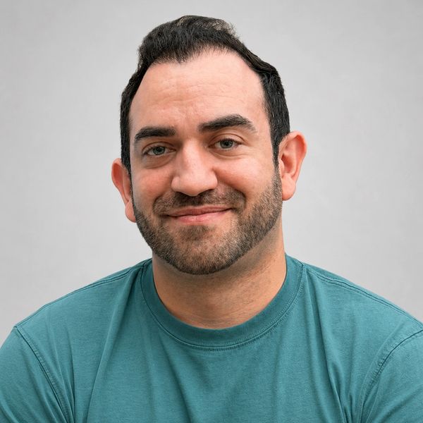 Smiling man with short dark hair and a teal shirt against a plain background.