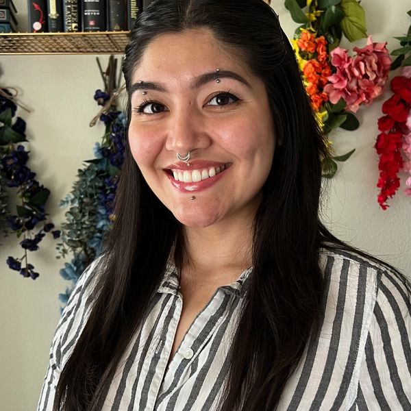 Smiling woman with piercings and long dark hair stands in front of colorful floral decorations.