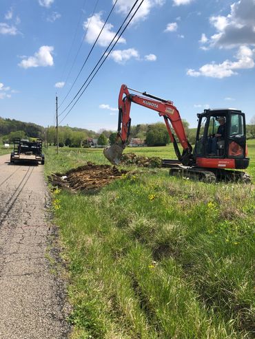 Excavator digging a trench beside a rural road on a sunny day.