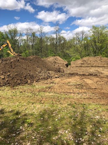 Excavator digging large piles of dirt near a wooded area under a cloudy sky.