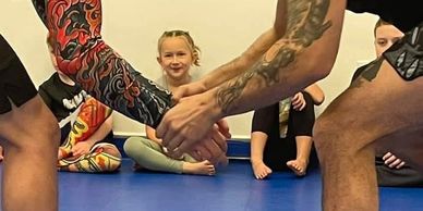 Two tattooed individuals practicing a martial arts grip with kids watching.