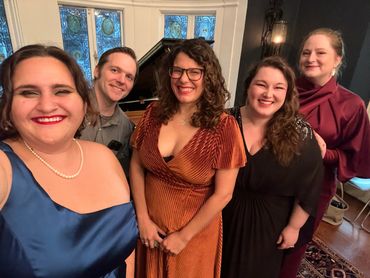A group of five friends dressed elegantly, smiling indoors near a piano.