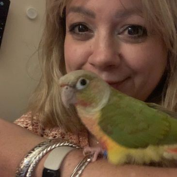 Woman smiling with her green and yellow pet bird perched on her arm.