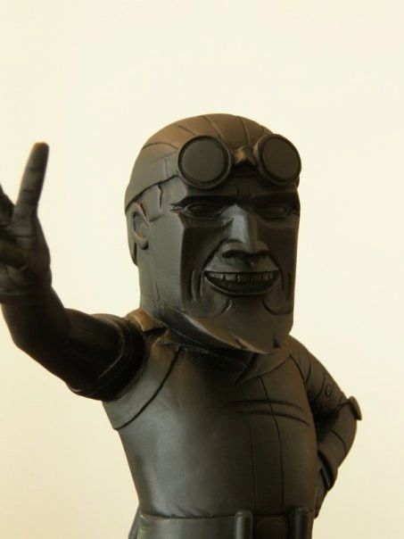 Black statue of a smiling figure with goggles and a raised hand gesture.