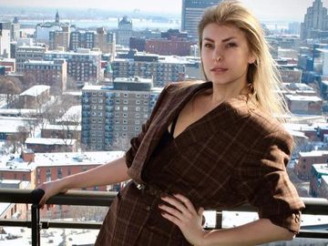 Femme en manteau marron sur balcon avec vue sur la ville enneigée.